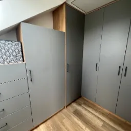 Szara szafa wnękowa z szufladami i czarnymi uchwytami, wkomponowana w skos dachu, z widoczną strukturą drewna na krawędziach i podłodze.