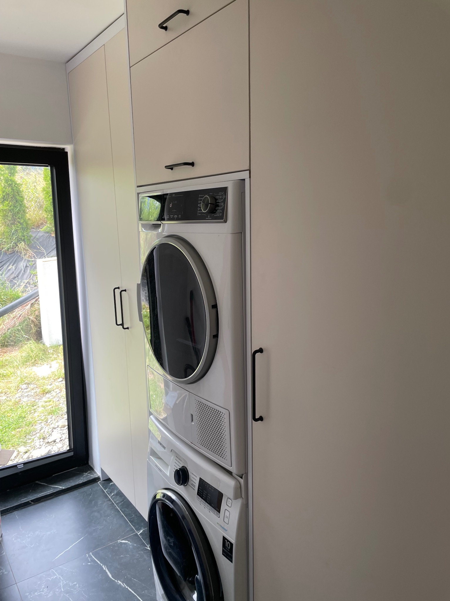 Jasna zabudowa meblowa z pralką i suszarką w pomieszczeniu z oknem i ciemną podłogą. Minimalistyczny design z czarnymi uchwytami.