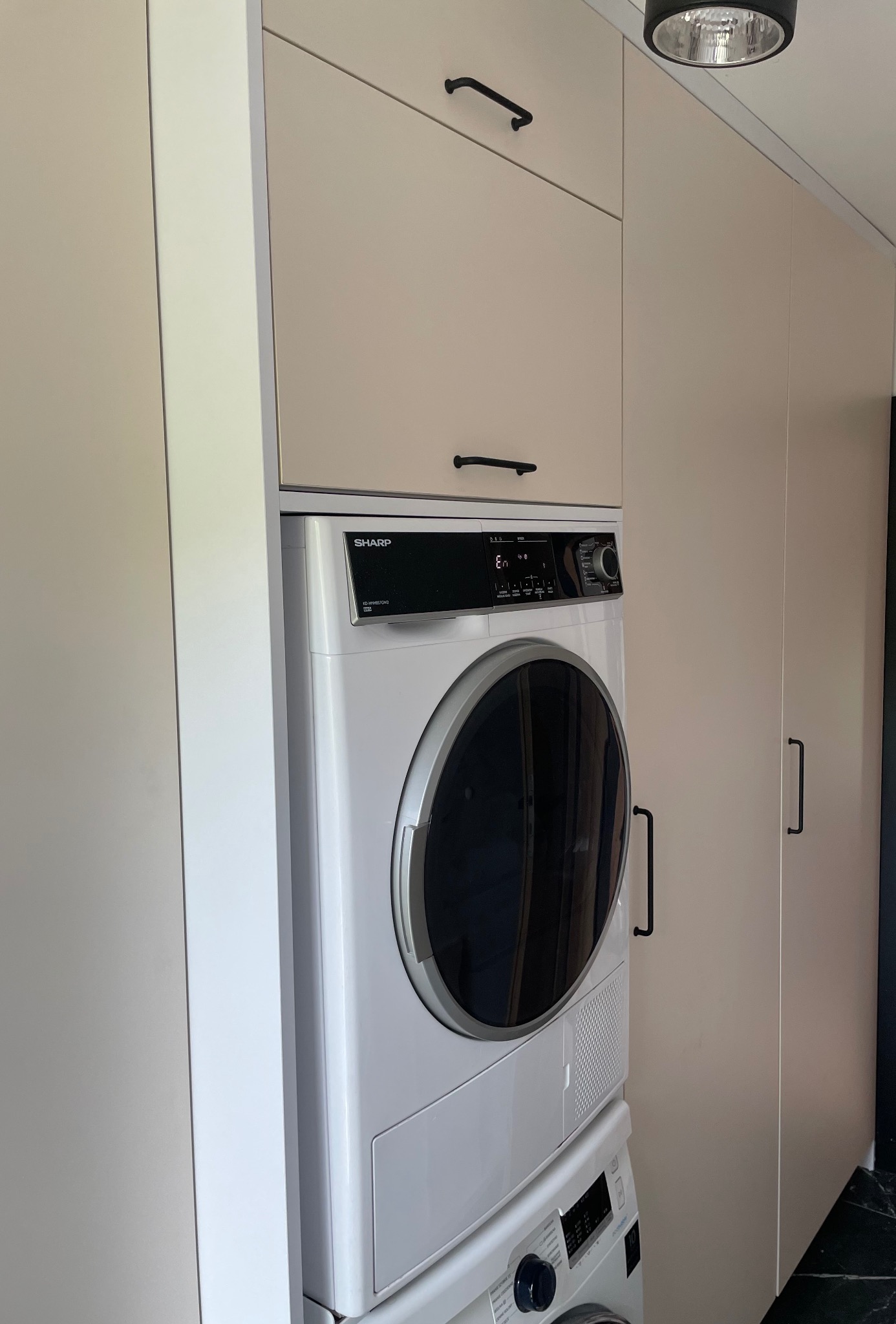 Pralka i suszarka w zabudowie meblowej z beżowymi frontami i czarnymi uchwytami, widoczne na tle szafy i czarnej podłogi.