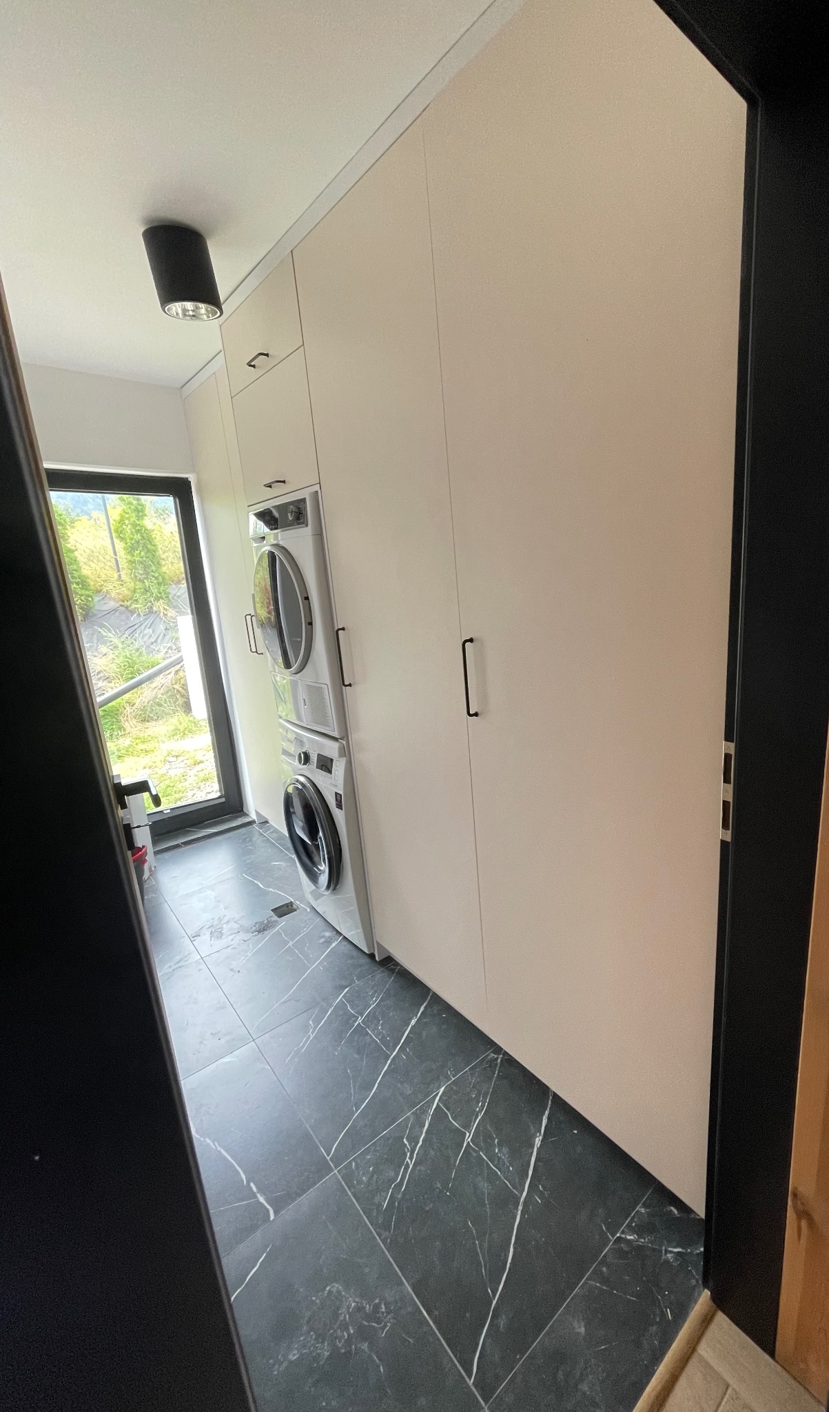 Biała szafa wnękowa z pralką i suszarką w nowoczesnej łazience z czarną marmurową podłogą, widok zza czarnej framugi drzwi.