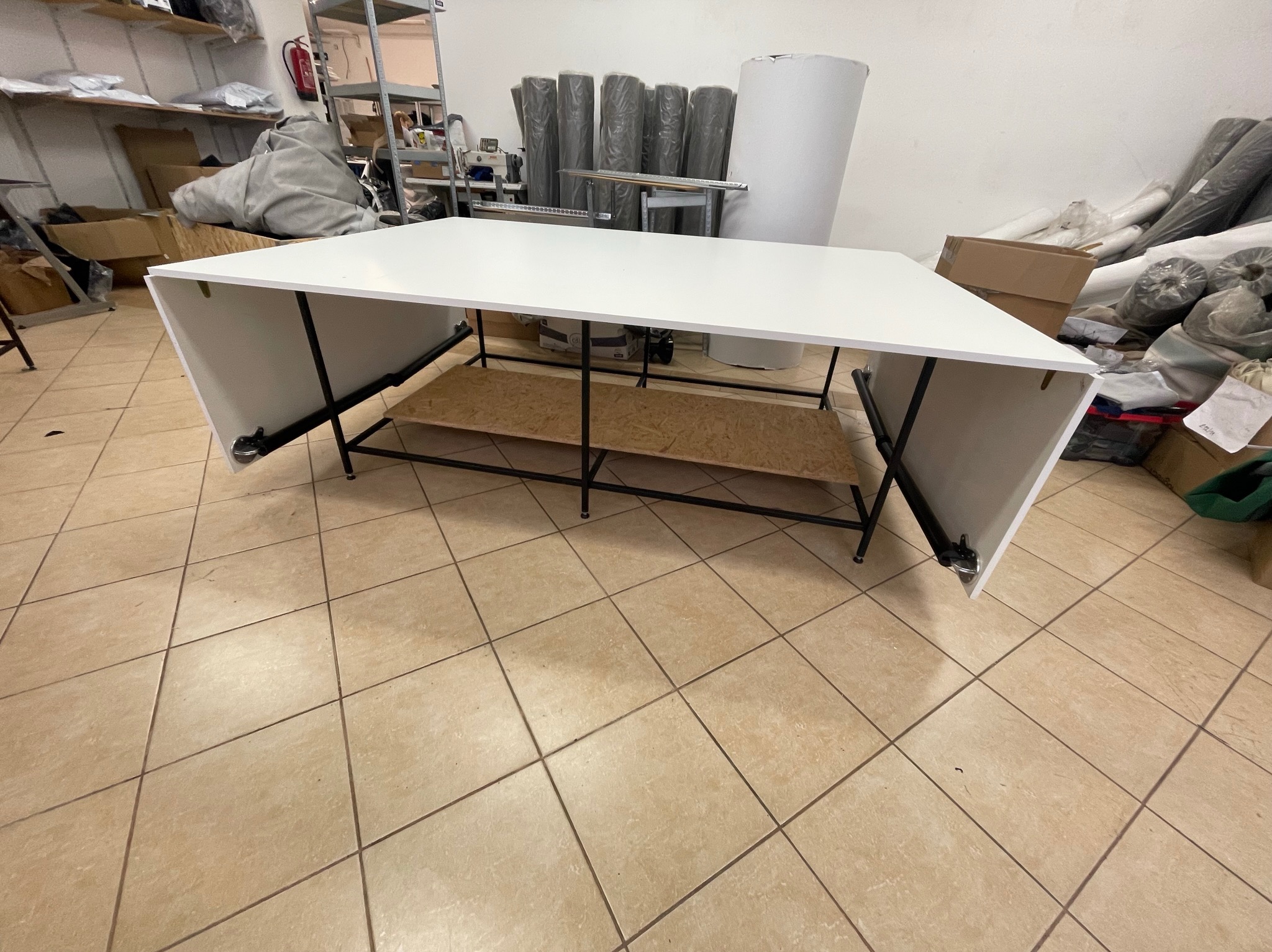 Stół krojczy z białym blatem i metalową konstrukcją, w tle rolki materiałów i maszyna do szycia. Widoczne płytki podłogowe i elementy warsztatu.