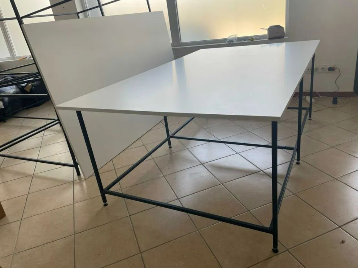 Biały stół z czarnymi nogami ustawiony na kafelkach, z białym panelem za nim, w tle widoczne okno i elementy wyposażenia.