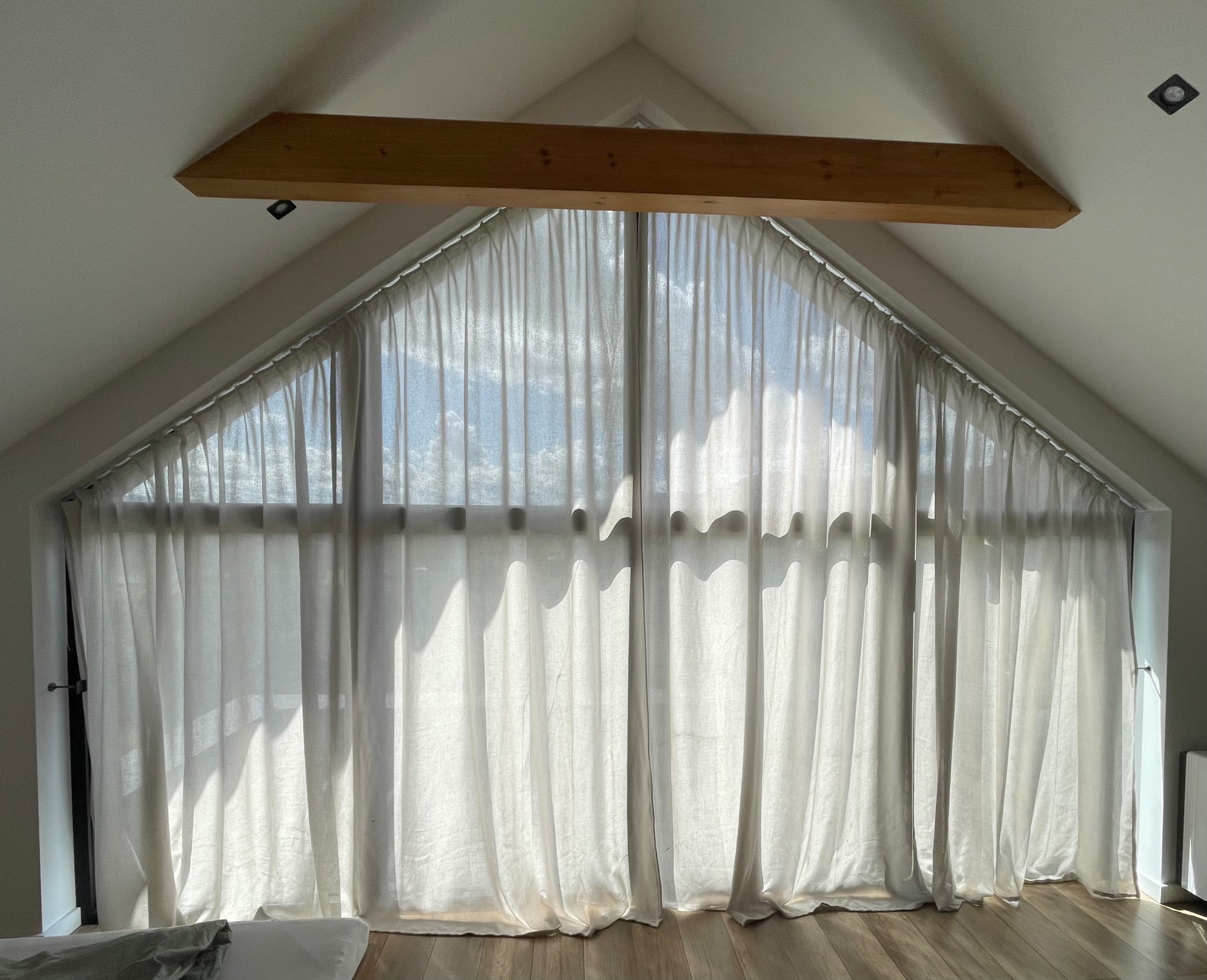 Jasne, przesuwne zasłony w oknie o nietypowym, trójkątnym kształcie, umieszczonym na poddaszu z drewnianą belką nad oknem. Widoczne promienie słoneczne.