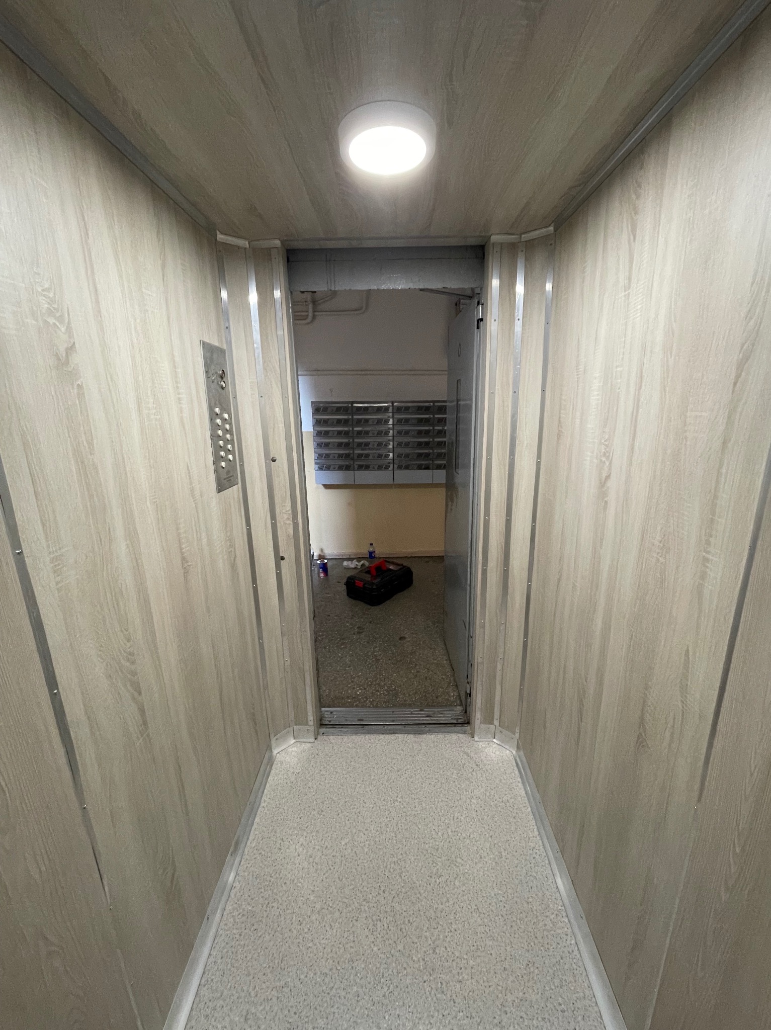 Wnętrze windy osobowej z panelami w kolorze jasnego drewna i metalowym panelem sterowania. Widok na otwarte drzwi i korytarz z kratką wentylacyjną.