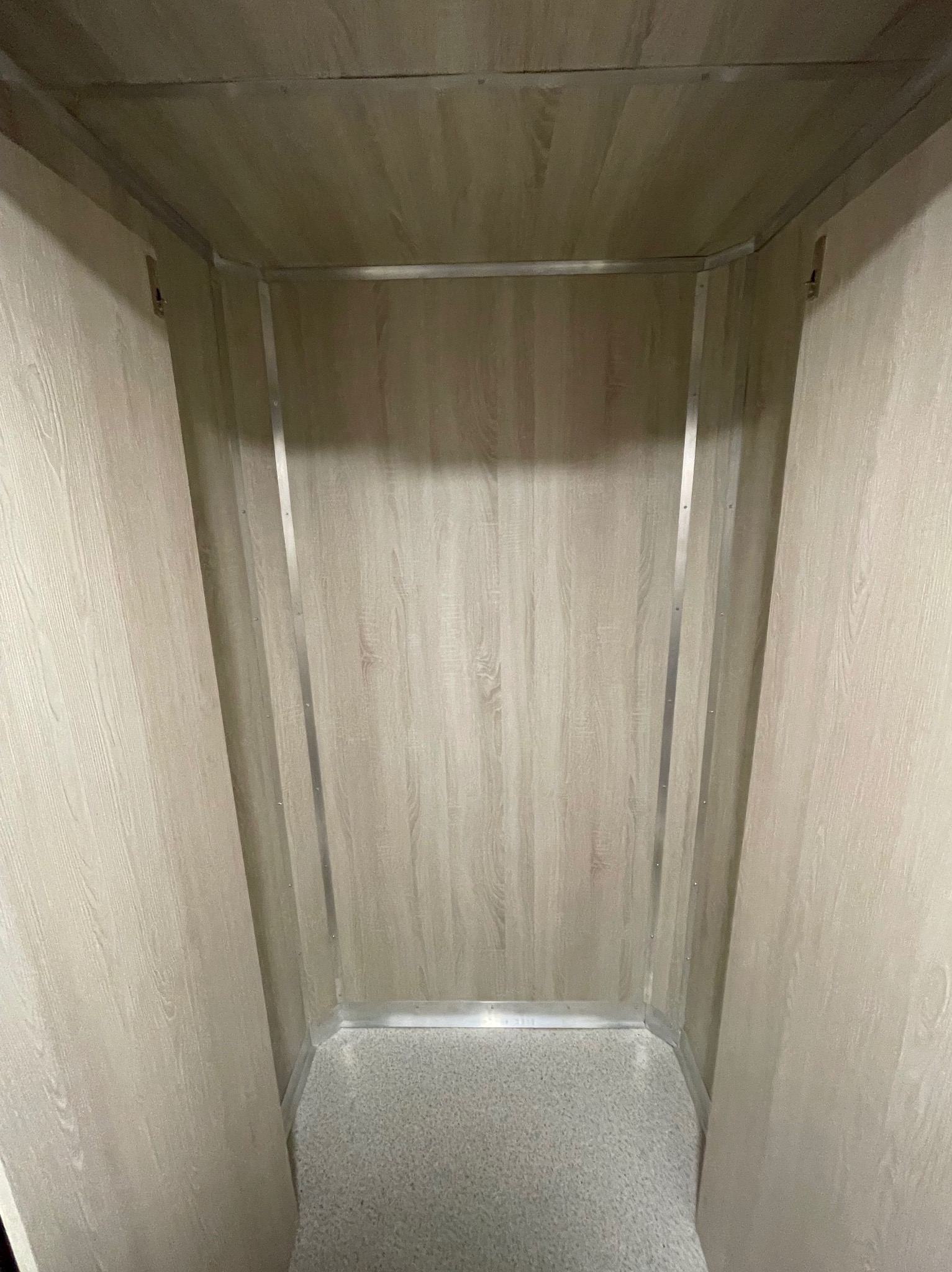 Wnętrze windy osobowej z panelami imitującymi drewno i metalowymi listwami wykończeniowymi. Widok z dołu kabiny, neutralna kolorystyka, symetryczny układ.