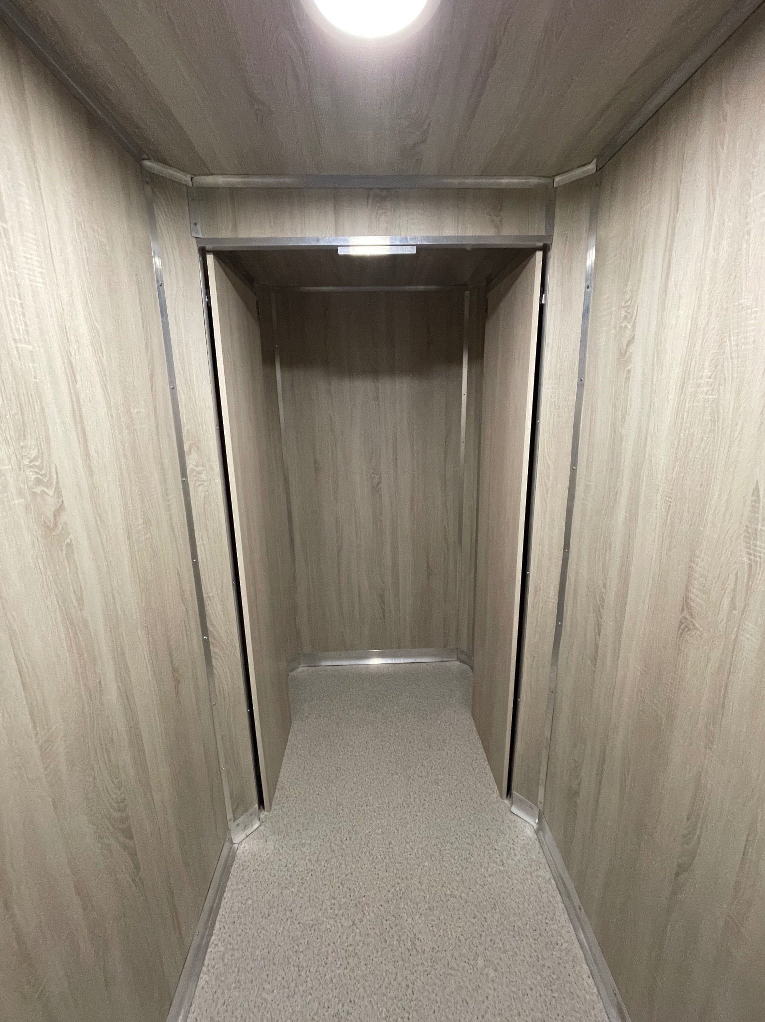 Wnętrze windy towarowej z panelami imitującymi drewno i szarą podłogą. Metalowe wykończenia krawędzi. Widok z perspektywy osoby wchodzącej.