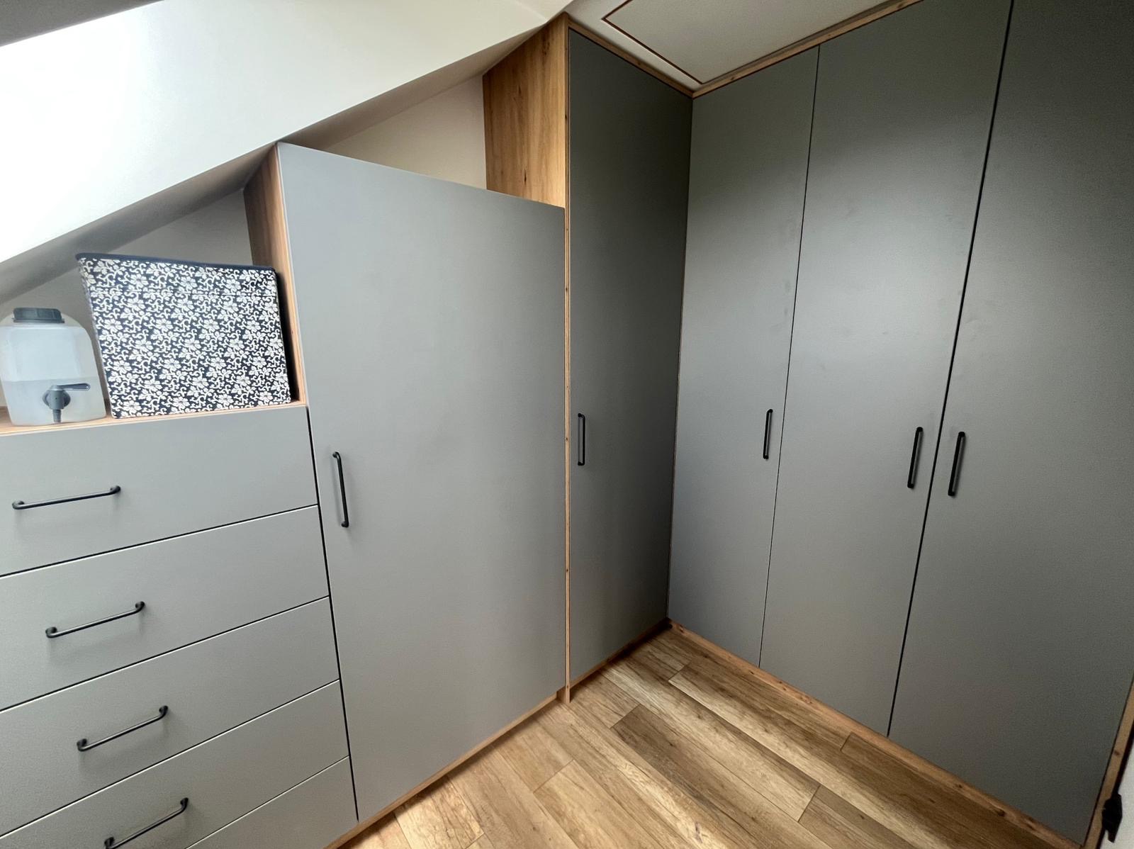 Szara szafa wnękowa z szufladami i czarnymi uchwytami, wkomponowana w skos dachu, z widoczną strukturą drewna na krawędziach i podłodze.