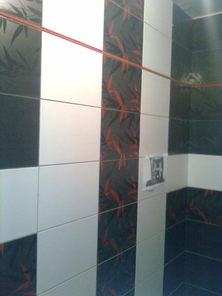 Ściana wyłożona czarno-białymi płytkami z motywem liści oraz czerwoną fugą, z widocznym fragmentem narożnika i kartką przyklejoną do płytek.