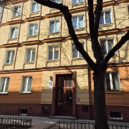 Fasada kamienicy z numerem 13, częściowo zasłonięta przez drzewo. Widoczne okna z białymi ramami, chodnik z kostki i metalowe ogrodzenie.