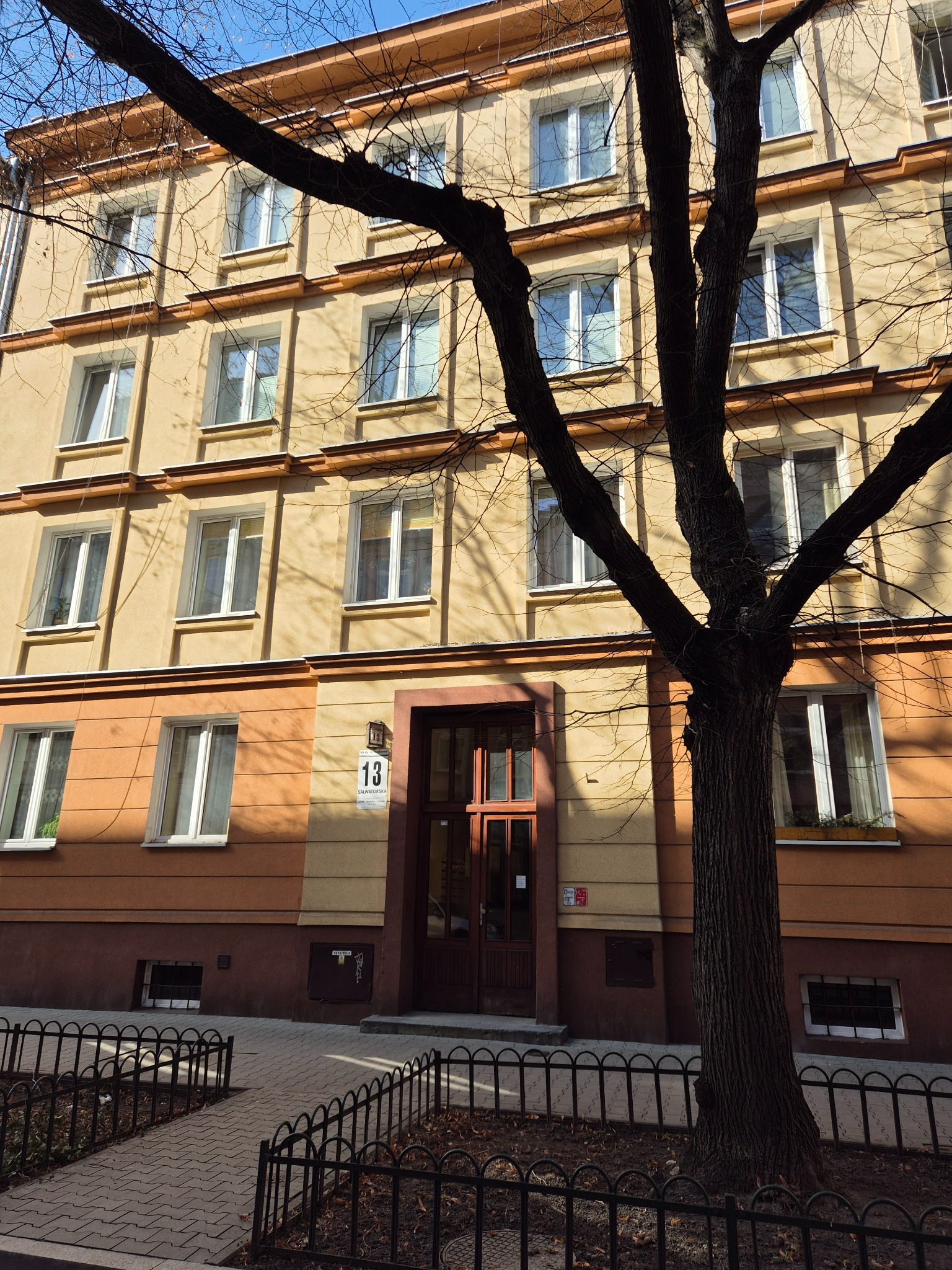 Fasada kamienicy z numerem 13, częściowo zasłonięta przez drzewo. Widoczne okna z białymi ramami, chodnik z kostki i metalowe ogrodzenie.