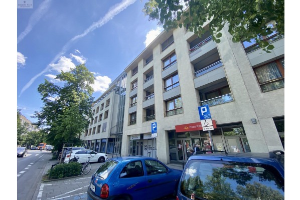 Budynek biurowy z balkonami, błękitne niebo w tle. Na pierwszym planie fragment niebieskiego samochodu. Widoczne znaki parkingowe i szyld Poczty Polskiej.