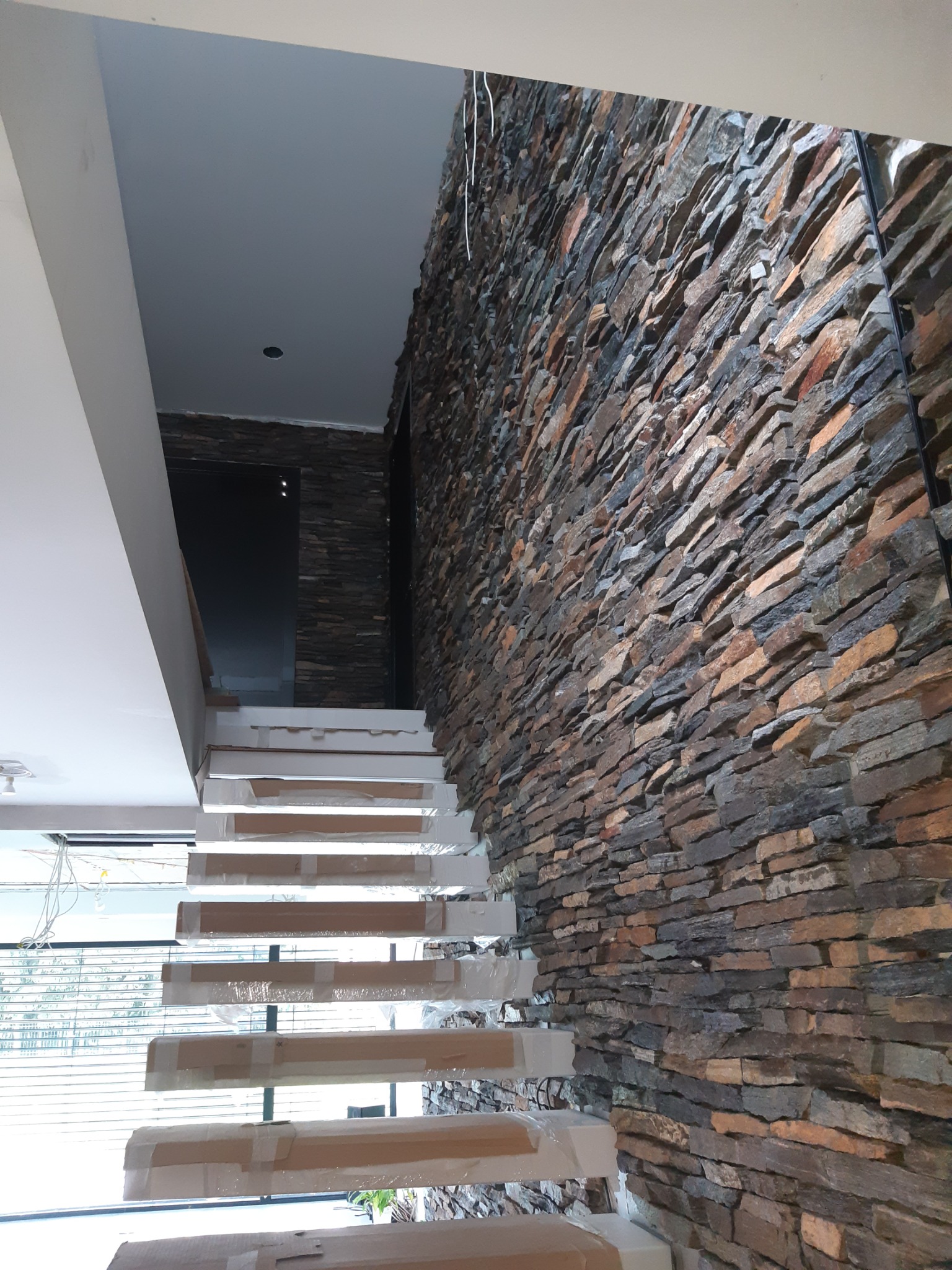 Ściana wyłożona nieregularnym kamieniem dekoracyjnym w odcieniach brązu i szarości, sąsiadująca z białymi schodami zabezpieczonymi folią ochronną podczas remontu.