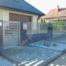 Nowoczesna, pozioma brama wjazdowa z szarego metalu, osadzona na niskim murku z ciemnej cegły. Widoczny fragment domu z numerem 23 i zadbany ogr&oacute;d.