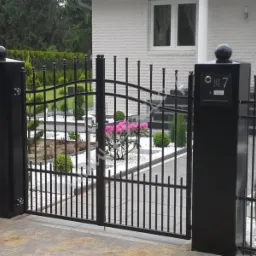 Elegancka, czarna, metalowa brama z numerem domu '7' na słupku, prowadząca do posesji z białego kamienia. W tle widoczne rośliny i zadbany ogród.