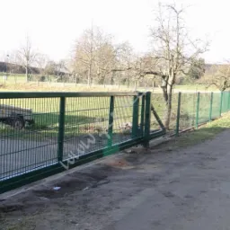 Zamykana, przesuwna brama panelowa w kolorze zielonym, z widocznym mechanizmem sterującym, na tle zielonego ogrodzenia panelowego i drzew.