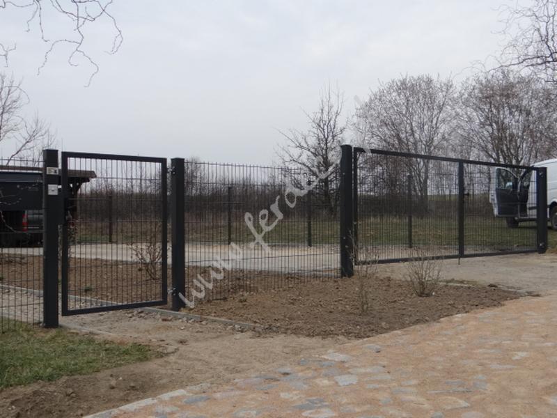 Nowoczesne ogrodzenie panelowe w kolorze grafitowym, z furtką i bramą, otoczone ziemią i kostką brukową. W tle widoczny samochód dostawczy.