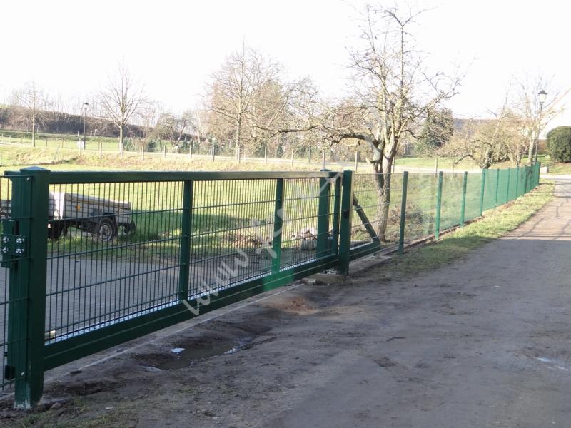 Zamykana, przesuwna brama panelowa w kolorze zielonym, z widocznym mechanizmem sterującym, na tle zielonego ogrodzenia panelowego i drzew.