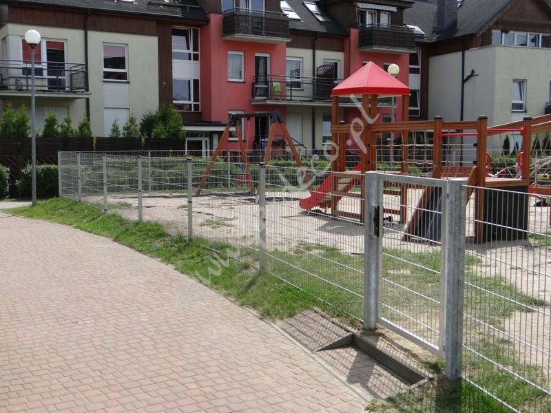 Srebrna siatka ogrodzeniowa z furtką oddzielająca chodnik od placu zabaw z kolorowym zestawem do zabawy na tle budynków mieszkalnych.