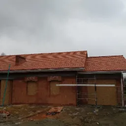 Surowy budynek jednorodzinny z czerwoną dachówką, częściowo osłoniętymi oknami i rusztowaniem w pochmurny dzień.