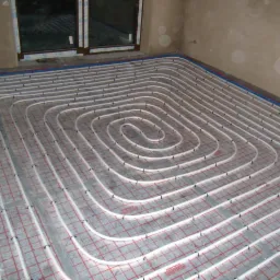 Białe rury ogrzewania podłogowego ułożone spiralnie na czerwonej siatce montażowej, widok z góry, surowy stan wykończenia wnętrza.