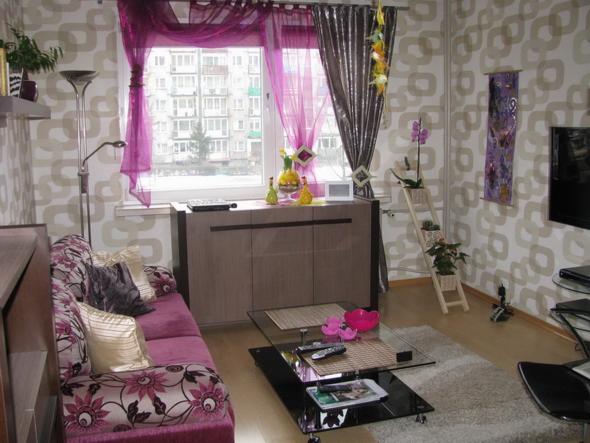 Jasne wnętrze pokoju dziennego z fioletowymi akcentami: sofa z kwiatowym wzorem, stolik kawowy ze szklanym blatem, szafka pod oknem z dekoracjami, okno z fioletowymi zasłonami, tapeta...