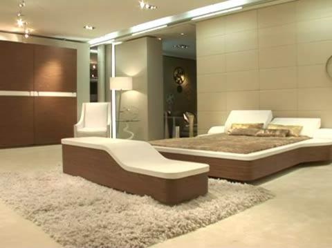 Nowoczesna sypialnia w beżach z łóżkiem na platformie, szezlongiem i puszystym dywanem, oświetlona światłem punktowym.