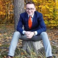 Uśmiechnięty mężczyzna w okularach, siedzący na pniu drzewa w jesiennym lesie, ubrany w niebieską marynarkę i pomarańczowy krawat.