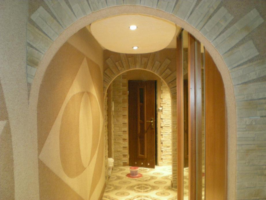 Wykończenie wnętrza z łukowatym przejściem, dekoracyjną ścianą z geometrycznym wzorem i kamienną okładziną, widoczne drzwi i oświetlenie sufitowe.