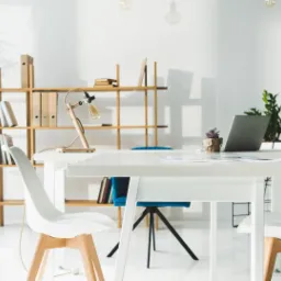 Jasne, minimalistyczne biuro z białym biurkiem, laptopem, lampką w stylu retro i roślinami doniczkowymi, widoczne krzesła i regał z dokumentami.