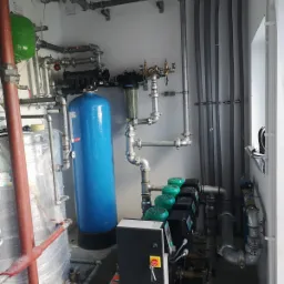 Kompleksowa instalacja w kotłowni: niebieski zbiornik ciśnieniowy, rozdzielacze z zielonymi pompami obiegowymi, liczne rury stalowe i plastikowe, oraz skrzynka sterownicza z kluczykami.