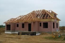 Dom w trakcie budowy: surowy stan z widoczną konstrukcją dachu z drewna, ściany z czerwonej cegły i częściowo otynkowane, drabina oparta o ścianę, teren wokół domu niezagospodarowany.