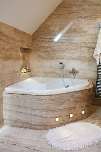 Narożna wanna obudowana trawertynem z podświetleniem LED w łazience wykończonej płytkami imitującymi kamień, widoczny okrągły dywanik i grzejnik drabinkowy.