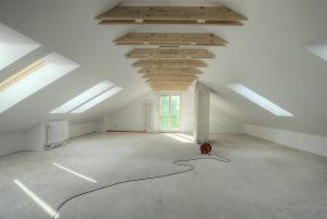 Poddasze po remoncie, przygotowane do dalszych prac wykończeniowych. Widoczne drewniane elementy konstrukcyjne dachu, okna dachowe i gładka, szara posadzka. Na podłodze leży zwinięty kabel...