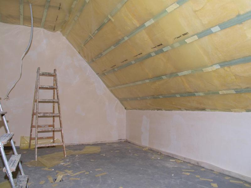 Poddasze w trakcie ocieplania, widoczne krokwie z izolacją z wełny mineralnej zabezpieczoną folią, ściany częściowo otynkowane, stara drabina oparta o ścianę, fragment drugiej drabiny z lewej strony.