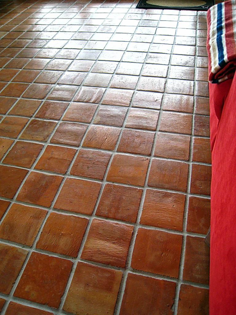 Ułożona pod kątem, rustykalna podłoga z terakoty w kwadratowe płytki z jasną fugą, widoczny fragment czerwonej sofy i dywanika.