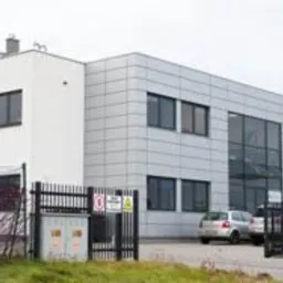 Nowoczesny, dwukondygnacyjny budynek przemysłowy z płaskim dachem, wykończony panelami w kolorze jasnoszarym i białym. Widoczne duże okna, metalowe ogrodzenie z tabliczkami ostrzegawczymi...