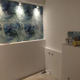 Wnętrze łazienki w trakcie prac wykończeniowych, widoczne płytki z motywem liści na ścianie, białe płytki z fakturą oraz ułożona w jodełkę podłoga z płytek imitujących drewno. Częściowo zamontowana...