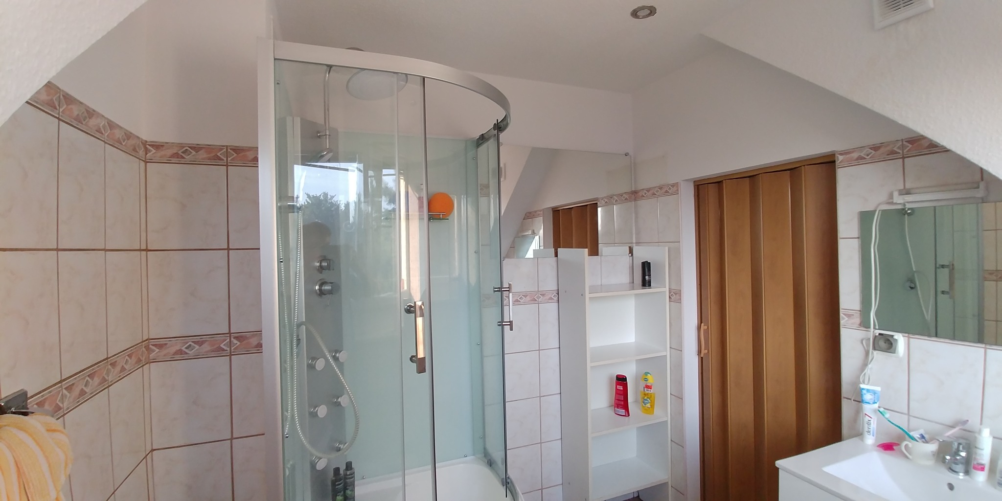 Zaokrąglona kabina prysznicowa z hydromasażem w łazience z beżowymi płytkami, widoczna półka z kosmetykami i składane drzwi.