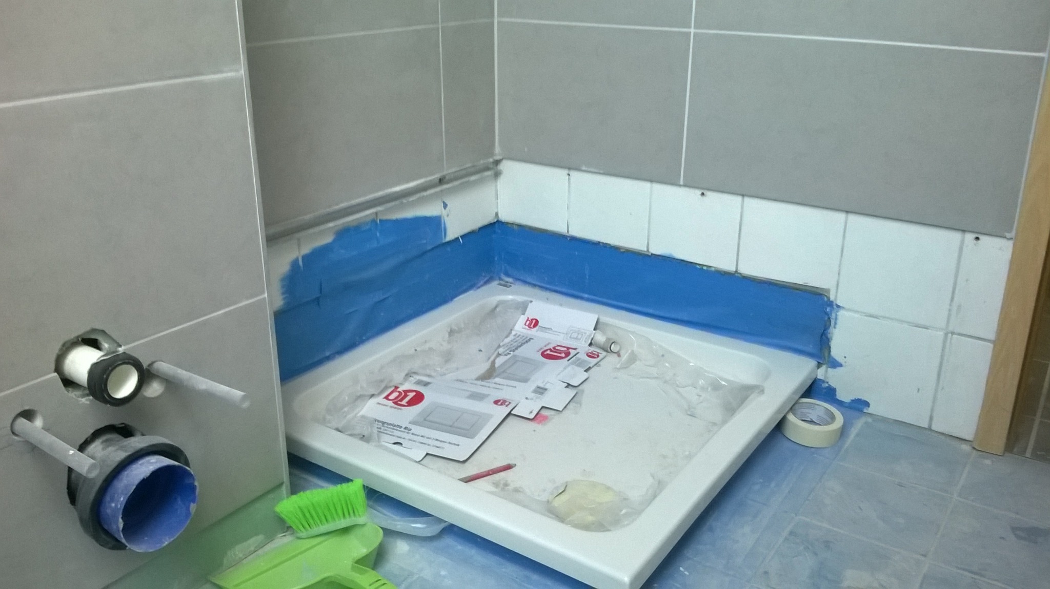 Instalacja brodzika w łazience: zabezpieczone ściany niebieską folią malarską, widoczne rury odpływowe w ścianie, instrukcje montażu brodzika wewnątrz niego, zielona szczotka do fug na podłodze.