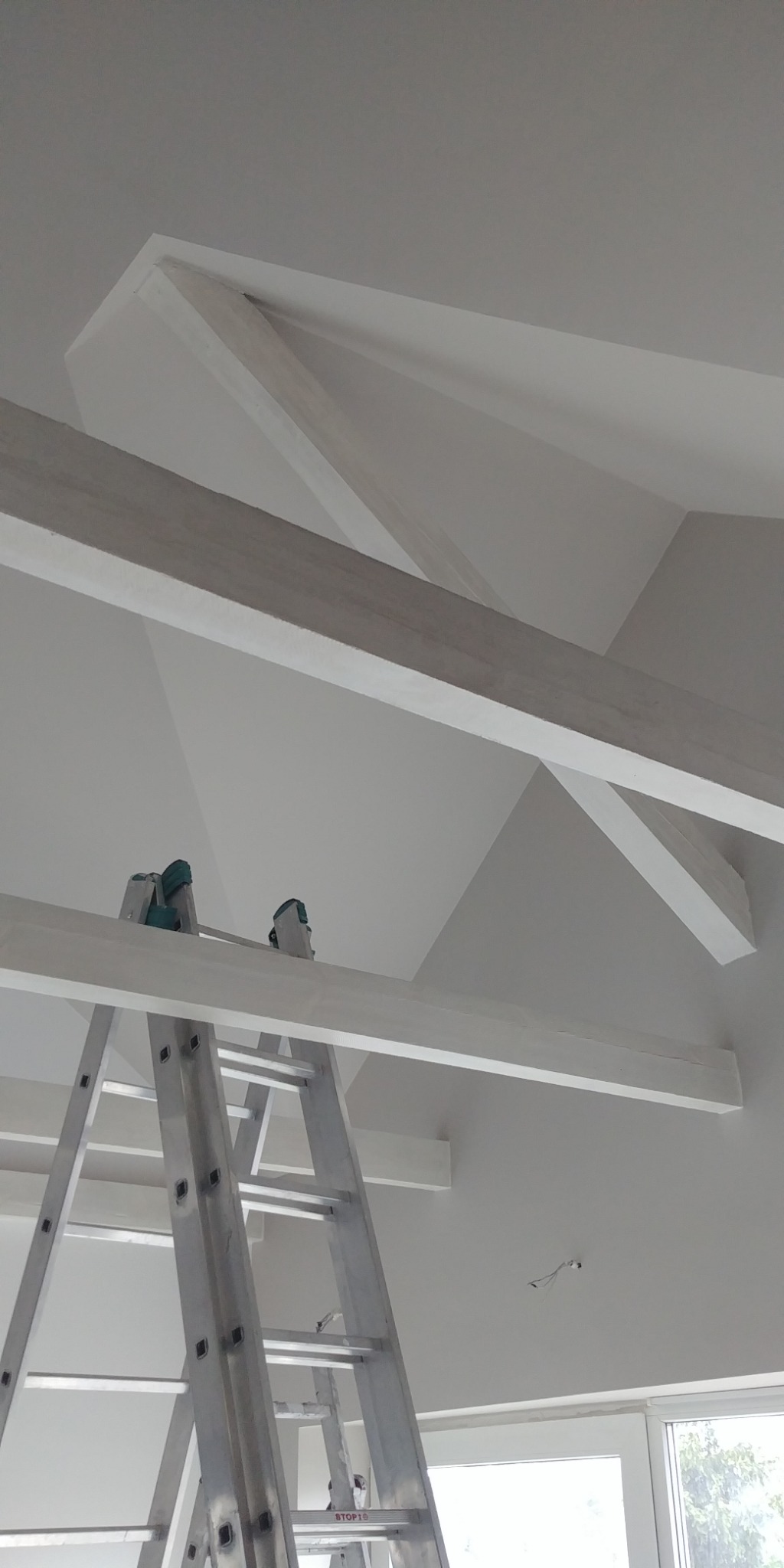 Białe, drewniane belki stropowe w nowym domu, widoczne z dołu z drabiny aluminiowej, z zielonymi ochraniaczami na szczycie, przy oknie.