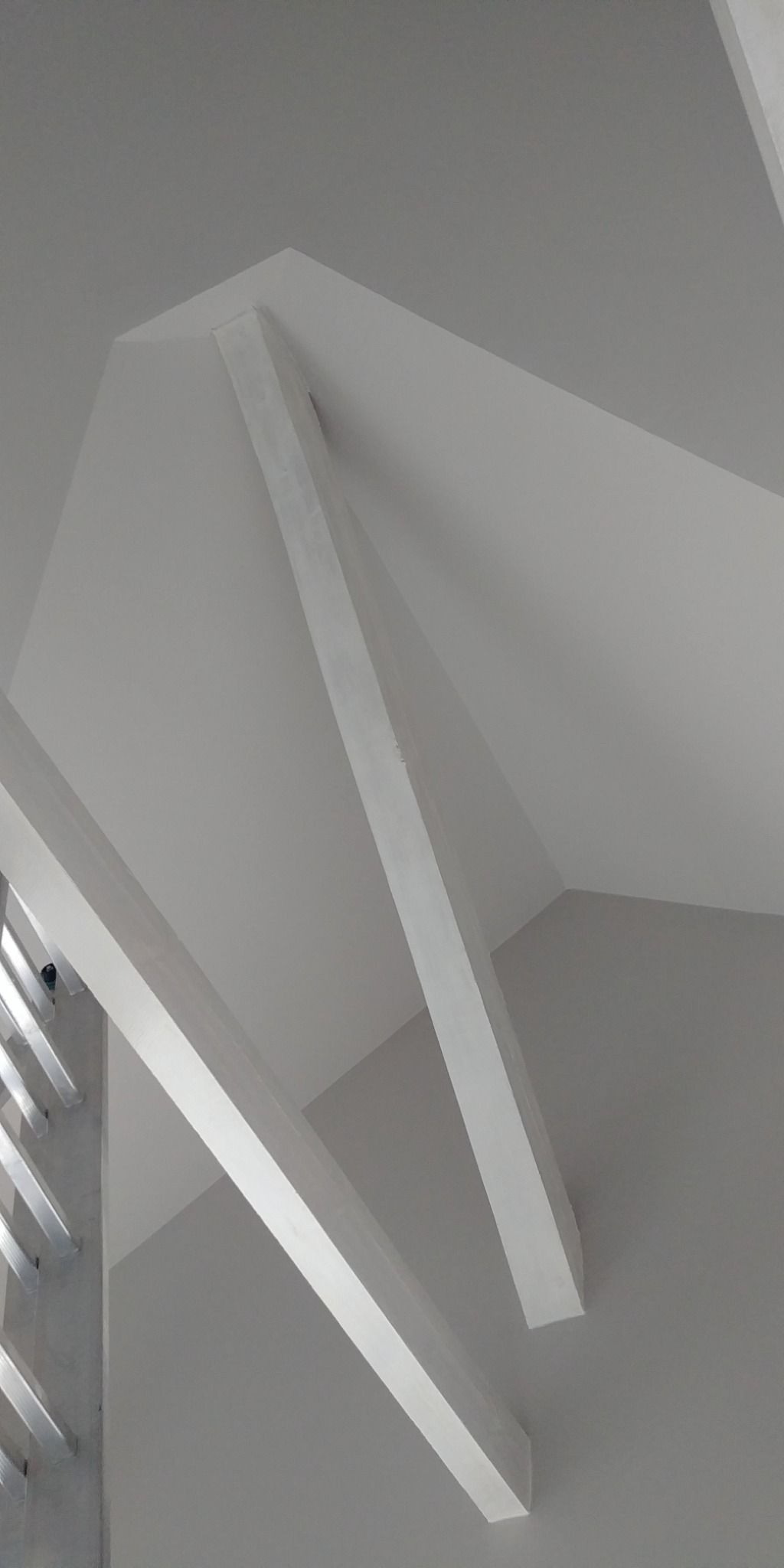 Białe, pomalowane belki stropowe pod skośnym sufitem, widoczne od dołu pod kątem, z fragmentem aluminiowej drabiny po lewej stronie.