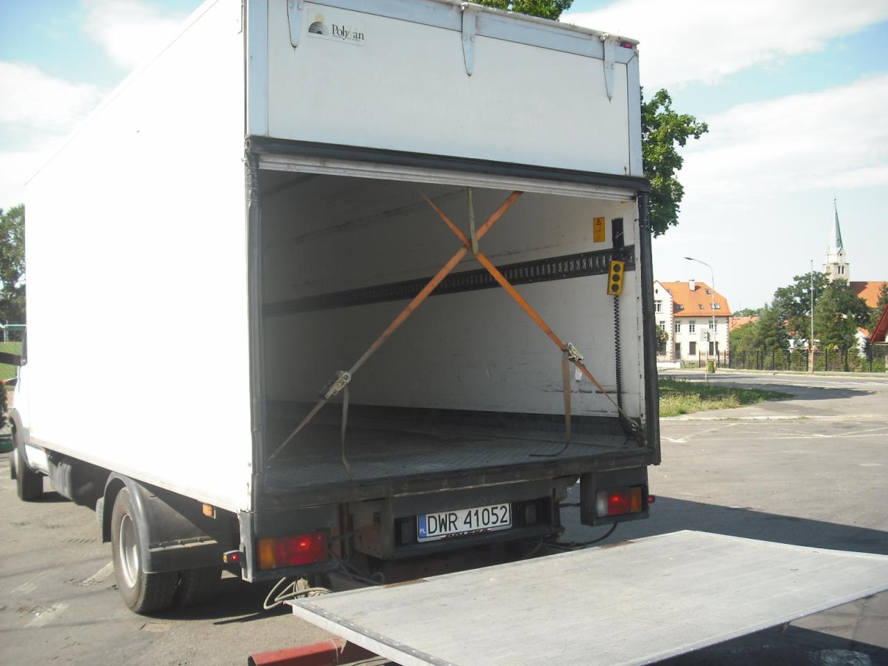 Otwarta paka białego busa dostawczego 3.5T z widocznymi pasami mocującymi ładunek, klapa załadunkowa opuszczona, w tle zabudowa miejska z wieżą kościoła.