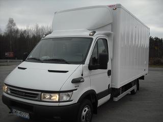 Biały samochód dostawczy Iveco z zabudową kontenerową, widoczny z przodu pod kątem, na szarym tle betonowego placu i zalesionego horyzontu.