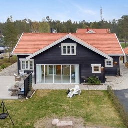 FLINK Scandinavian Houses Spółka z o.o. - Czarny dom z pomarańczową dachówką i przeszkloną ścianą frontową, widok z góry na taras z meblami ogrodowymi i trawnik z grillem trójnożnym.