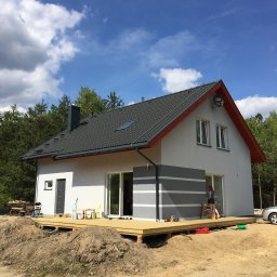 FLINK Scandinavian Houses Spółka z o.o. - Nowoczesny dom jednorodzinny z szarą dachówką, drewnianym tarasem na podwyższeniu i elementami dekoracyjnymi w postaci szarych pasów na elewacji, w tle las i błękitne niebo z chmurami.