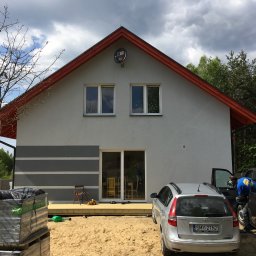 FLINK Scandinavian Houses Spółka z o.o. - Nowoczesny dom z szarą elewacją, czerwonym dachem i satelitą na szczycie, z drewnianym tarasem w trakcie budowy, zabawki na tarasie, palety z materiałami budowlanymi i srebrny samochód...