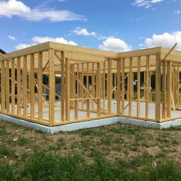 FLINK Scandinavian Houses Spółka z o.o. - Surowa drewniana konstrukcja domu w trakcie budowy, widoczny fundament z izolacją, w tle pracownik budowlany i błękitne niebo z chmurami.