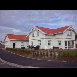 FLINK Scandinavian Houses Spółka z o.o. - Nowoczesny biały dom z czerwonym dachem i dużymi oknami tarasowymi, widok z zewnątrz, z widocznym tarasem i meblami ogrodowymi oraz przyległym budynkiem gospodarczym z otwartymi drzwiami.