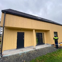 FLINK Scandinavian Houses Spółka z o.o. - Nowoczesny, parterowy budynek z jasną, drewnianą elewacją pionową, czarnym dachem i stolarką okienną w kolorze antracytowym, na tle pochmurnego nieba, z pracownikiem w odblaskowej kamizelce przy...