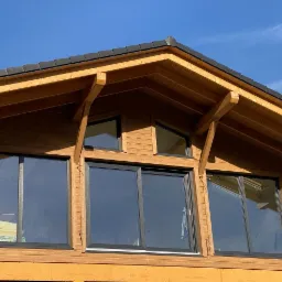 Fragment ściany domu szkieletowego z widocznymi trzema oknami i elementami konstrukcyjnymi z drewna, na tle błękitnego nieba.